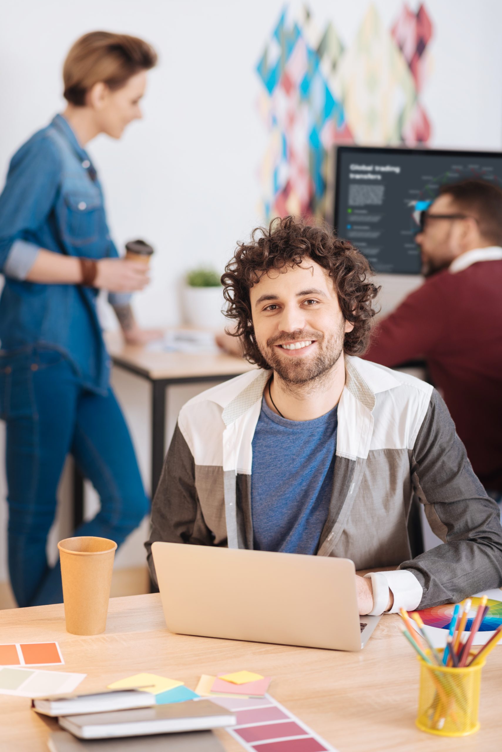 Pleased with work. Handsome joyful bearded man working on his laptop while sitting at the table and his colleagues working in the background