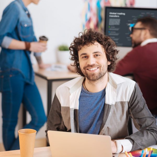 Inspired young man working on his laptop Pleased with work. Handsome joyful bearded man working on his laptop while sitting at the table and his colleagues working in the background